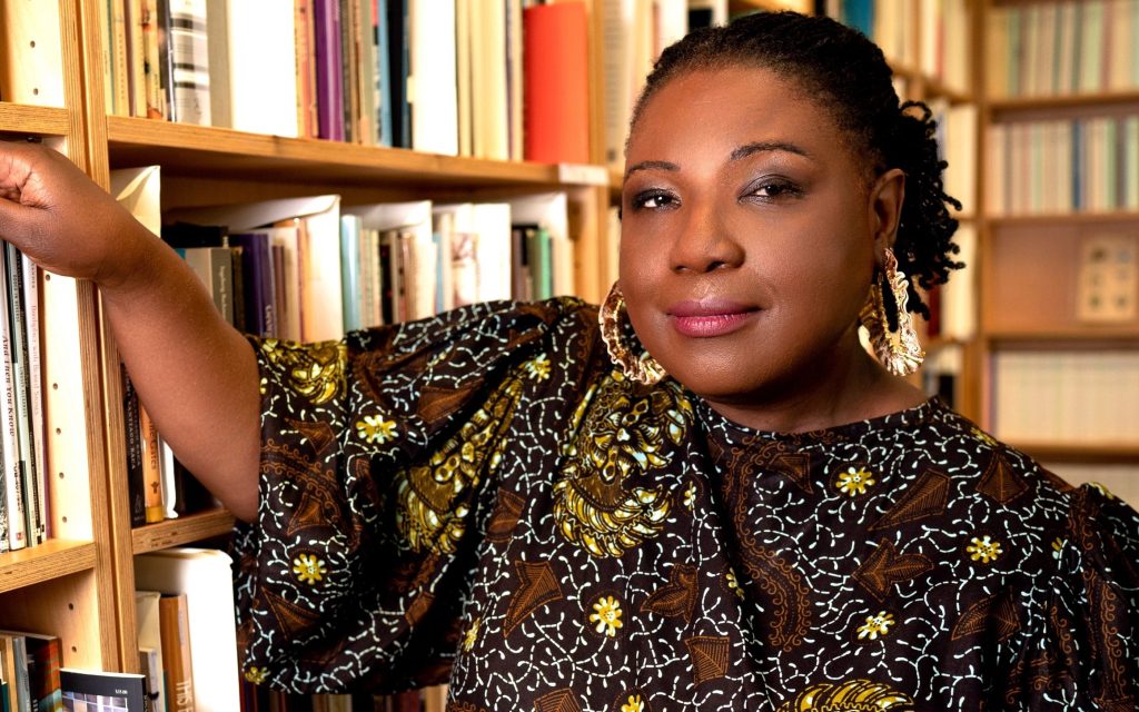 Library. Image is of a Black woman in a black , brown and white patterned dress leaning on a library bookshelf filled with books.
