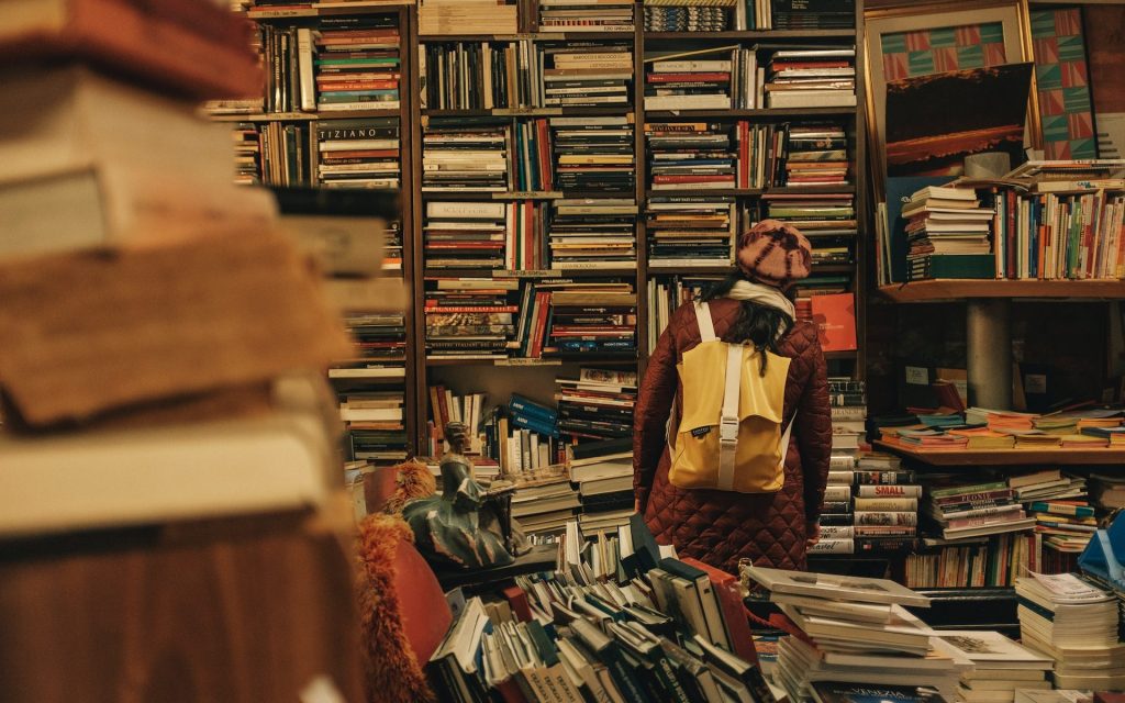 City of Literature. UNESCO. Image is a library or bookshop stuffed with books and a person wearing a backpack and beanie with their back to the camera looking at the titles.