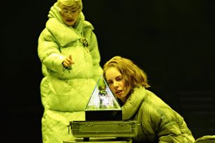 Oil. Image is of a older grey haired woman in glasses and bulky padded yellow/green coat, who is pointing at a prism, which is also being looked at by a younger woman in similar attire with shortish brown hair and kneeling down.