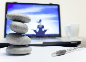 Zen rock stack in foreground with a computer screen showing silhouette of woman in yoga pose balancing social media with screen.
