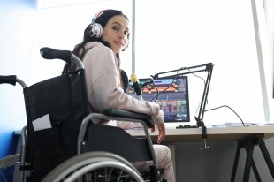 Accessibility. Image shows young woman with long dark hair and a light top in front of computer screen and microphone. She is looking at the camera over her shoulder. She is wearing headphones and using a wheelchair.