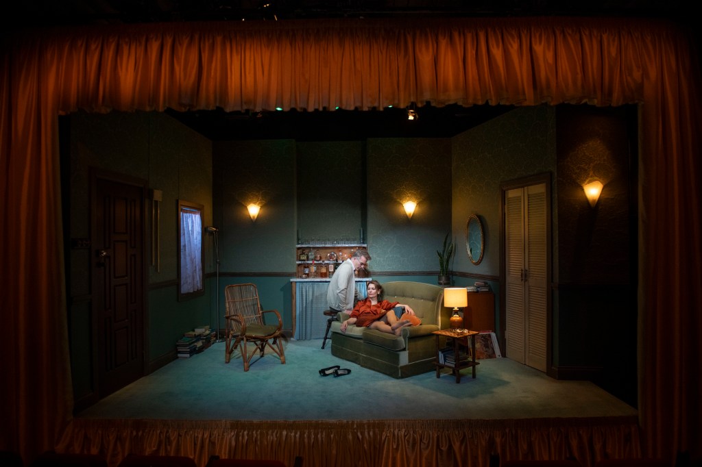 Who's Afraid of Virginia Woolf. Image is a proscenium arch stage set of a dingy living room, with a woman in a red shirt draped across the couch and a man in a grey cardigan sitting on the edge leaning toward her.