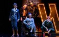 The Master and Margarita. Image is a group of five actors sitting, crouching or standing on stage in front of a huge lit W sign.