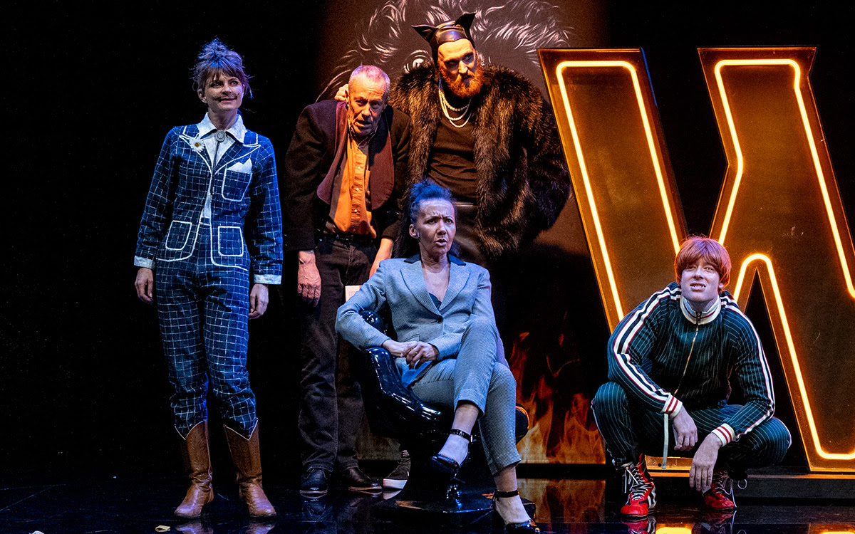 The Master and Margarita. Image is a group of five actors sitting, crouching or standing on stage in front of a huge lit W sign.