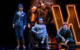 The Master and Margarita. Image is a group of five actors sitting, crouching or standing on stage in front of a huge lit W sign.