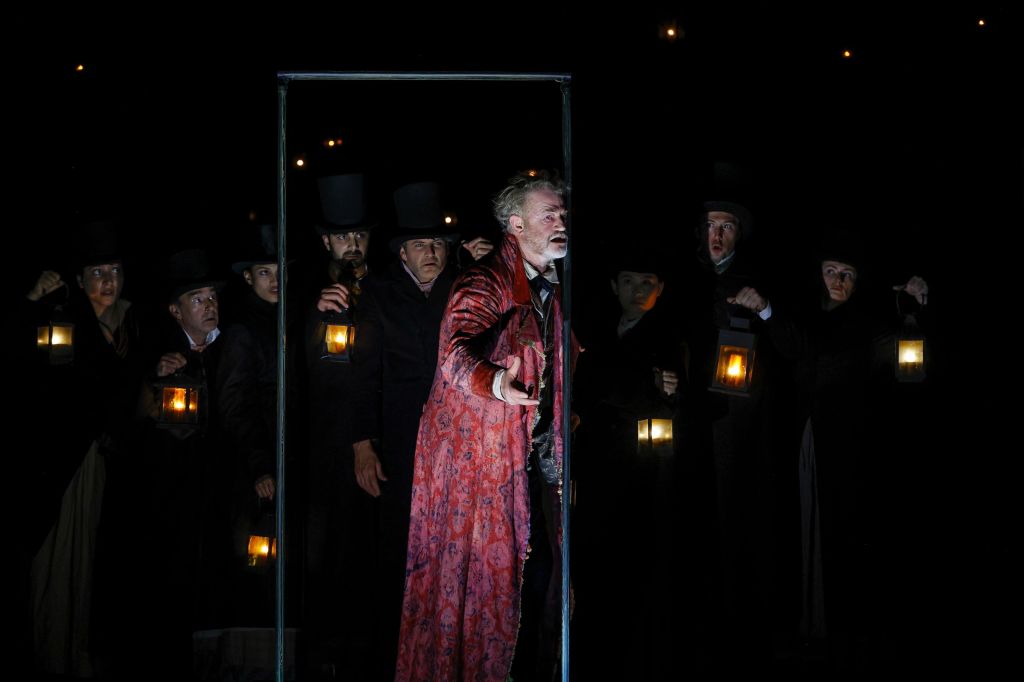 A Christmas Carol. Image is a darkened stage with an ensemble of Dickensian actors holding lanterns and Scrooge in a reddish dressing gown looking out through a door frame.
