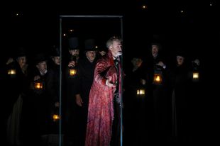 A Christmas Carol. Image is a darkened stage with an ensemble of Dickensian actors holding lanterns and Scrooge in a reddish dressing gown looking out through a door frame.