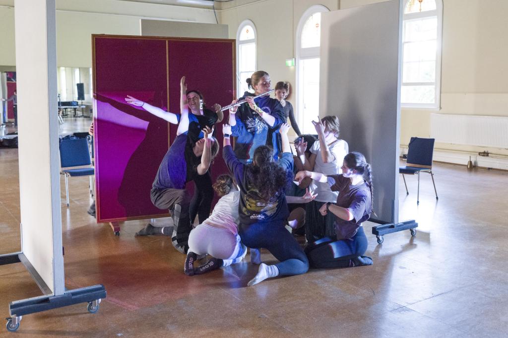 a group of young performing arts students clustered around a girl playing a flute.