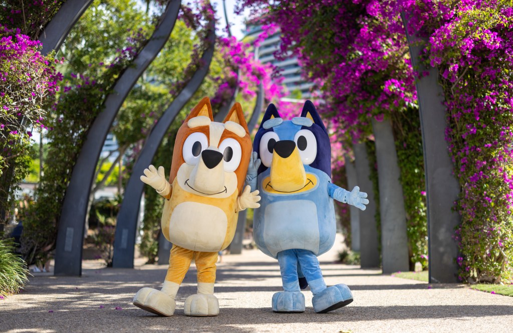 Bluey's World. Image is animation representation of a floral walkway in Brisbane, with a brown and yellow dog figure and a blue one waving at the camera.
