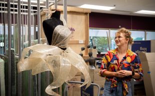 Finalist in 'Exit Art' exhibition 2023, Jasper Mules from The Essington School, NT. Photo: Supplied. Inside an artwork stockroom a figure with curly blond hair looks towards a 3D fashion garment on the mannequin. The garment appears to be a short dress made out of translucent, petal-like materials that is cream in colour. The figure is smiling, wearing glasses and a colourful patterned shirt with jeans.