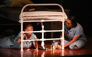 'Is That You, Ruthie?'. Two First Nations women are on stage in a dormitory, one sitting on the floor, one crouching underneath a metal framed bed. They are wearing grey tunics and playing with small wooden figurines.