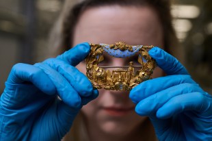 close-up of gold miner's brooch held by blue-gloved hands and woman's eyes peering through.