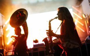 Upcoming free programming. Xenos, Mofo Sessions at Royal Park, Mona Foma 2021. Photo: Mona/Jesse Hunniford. Image: Courtesy of the artist and Mona. Two musicians backlit on the stage, the one on the left is playing a sousaphone and the one on the right a saxophone.