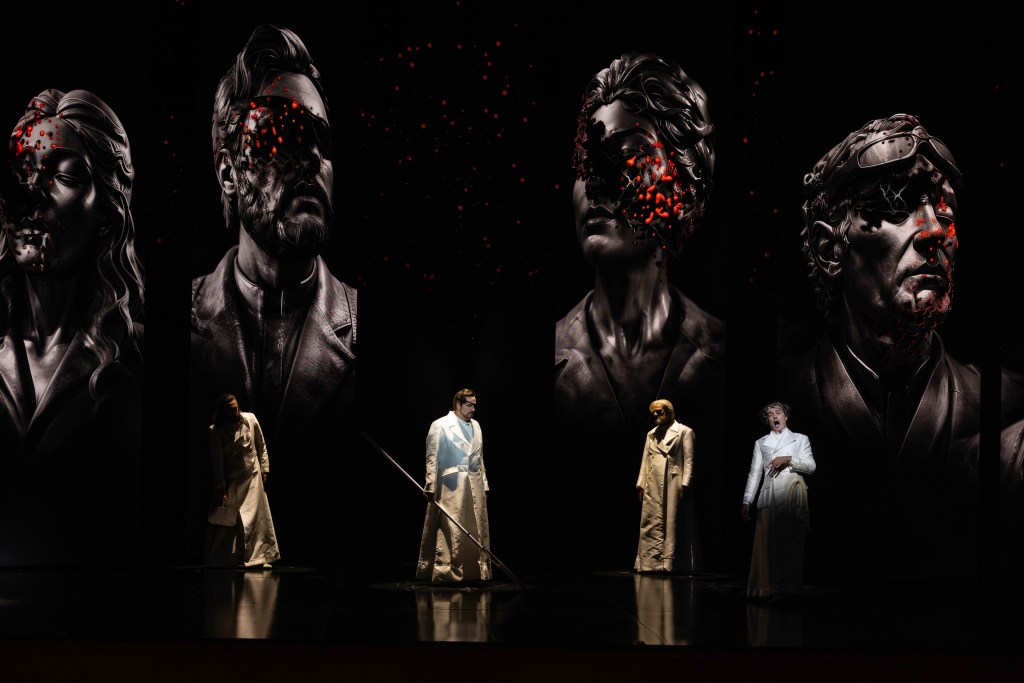 Das Rheingold. Four giants dark brown heads with splashes of red form the backdrop of the stage, on which stand four white robed opera singers playing Norse gods.