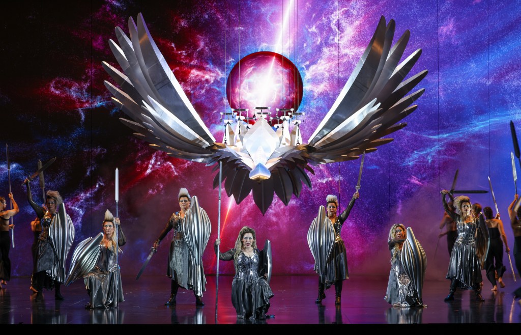 Die Walküre. A purple and reddish backdrop with a huge silver flying bird like figure above a group of silver clad warrior women.