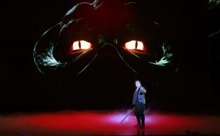 Siegfried. Lone man with sword on a red stage in front of huge image of glaring eyes.