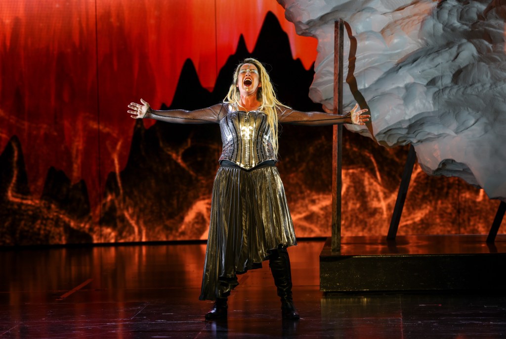 Götterdämmerung. A Wagnerian female opera singer with long blonde hair and body armour stands in the middle of a stage in front of a red fiery backdrop has her hands wide out to the side.