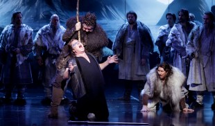 Götterdämmerung. A Wagnerian male singer is on his knees screaming out in distress, while a man behind him attacks him with a staff. Other figures stands out around in furs.