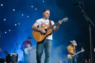 Zach Bryan. Image is a man in jeans and Budweiser T shirt playing an acoustic guitar in front of a band on stage. His right arm is heavily tattooed.