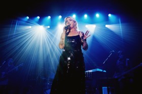 Always Live. Image is a blue lit stage with a blonde woman in a sleeveless dress singing or speaking into a microphone.