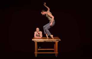 Two male circus performers, one leaning on a table, the other balancing on top of it.