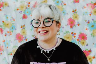 A photo of Claire Christian, a romance author with short, bleached hair and black glasses. She's standing against a flower background.