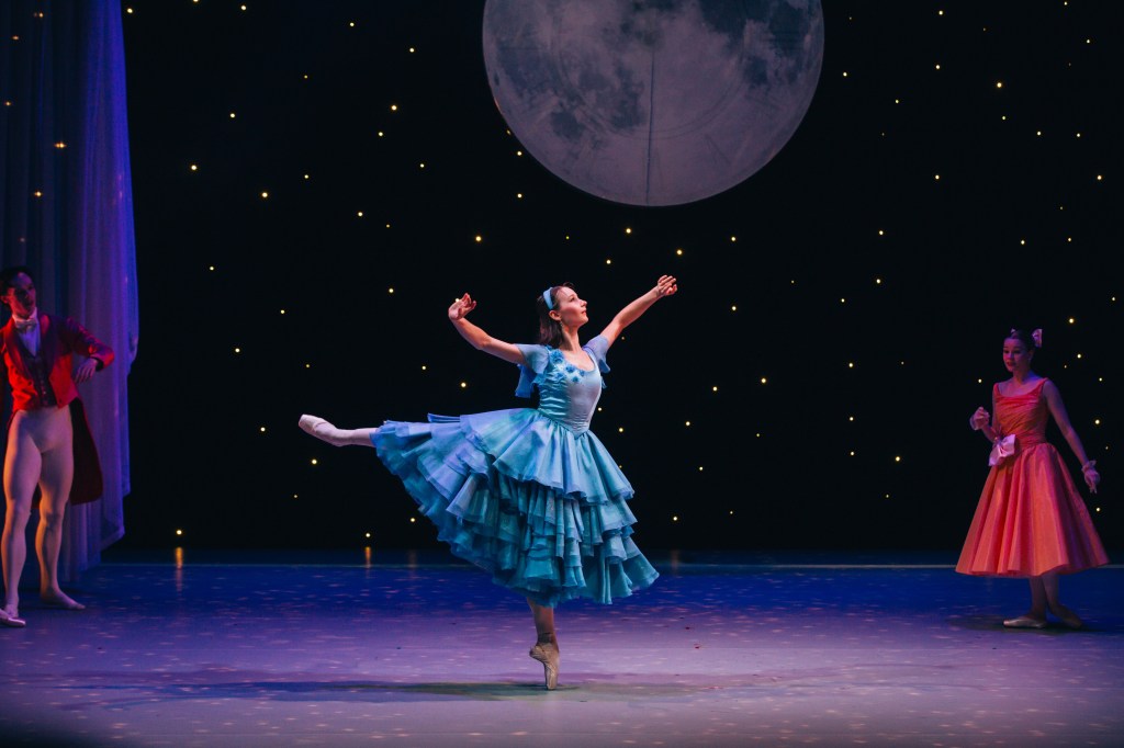Cinderella. Image is a ballet dancer in a blue layered costume dancing with arms out to the side and balanced en pointe with one leg raised behind her. She is on a stage with a large moon and starry sky as the backdrop. A male and female dancer are on the left and right hand side of the stage respectively.