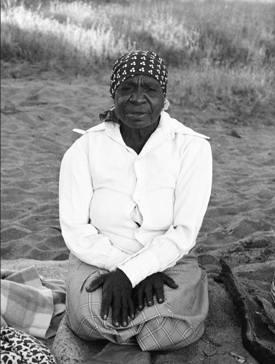 A slightly grainy black and white pphoto of Emily Kam Kngwarray, an elderly Aboriginal woman, kneeling with hands crossed on her knees, on a sandy landscape. 