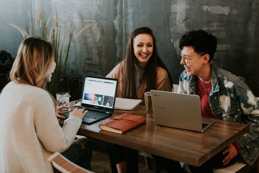 office work. A group of three young people (in their twenties) sitting around a table with their laptops, laughing,