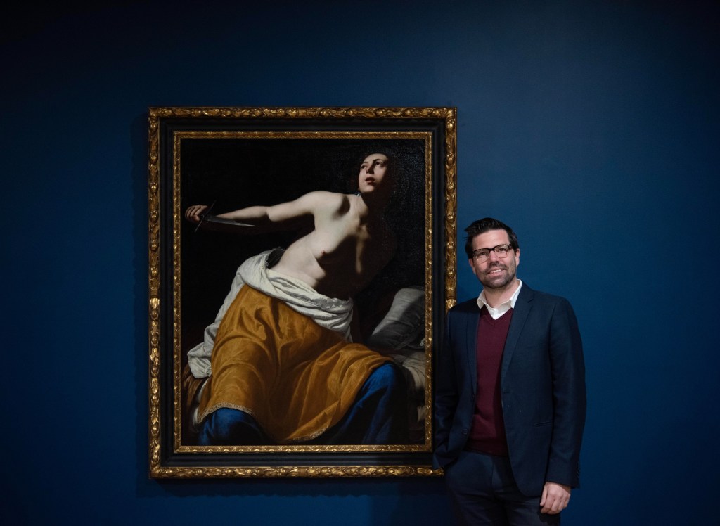 Hamilton Gallery Director Joshua White next to 'Lucretia' by Artemisia Gentileschi, installation view in Emerging From Darkness: Faith, Emotion and The Body in the Baroque at Hamilton Gallery. Photo: Madi Whyte. A figure with short brown hair, glasses and brown beard wearing a collared shirt and blazer stands next to a large painting depicting a women, half nude, sitting at the edge of her bed and gazing up.