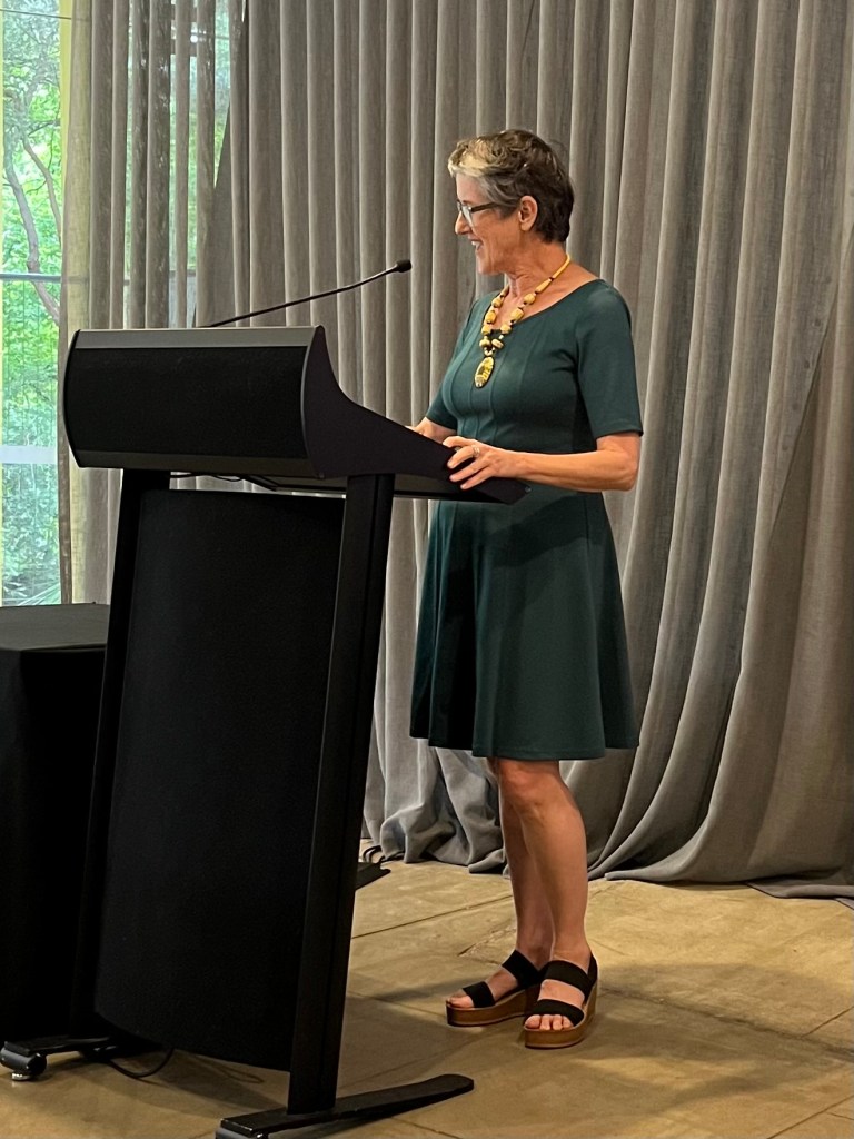 Sue Giles. A woman in a green dress stands at a lecture in front of a curtain speaking into a microphone, facing to the left of the shot. She is wearing glasses and a necklace.