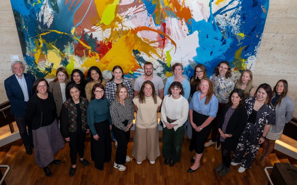 Arts Leadership Program. Image is a large group of people standing in front of a colourful backdrop and looking at the camera.