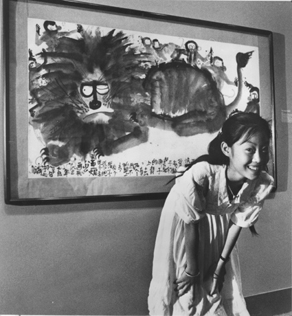 Black and white photo of a young figure with black hair in a pony-tail smiling and looking at the viewer. Behind the figure is a large ink drawing of a lion with one eye open.