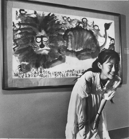 Black and white photo of a young figure with black hair in a pony-tail smiling and looking at the viewer. Behind the figure is a large ink drawing of a lion with one eye open. 