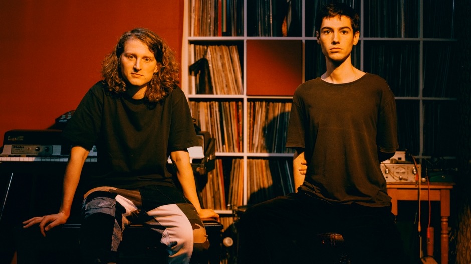 Two figures in an indoor setting with a large record rack behind them. The figure on the left has shoulder-length curly dark blond hair and is wearing a black tshirt with coloured pants. The figure on the right is with brown short hair and a black tshirt. Both of them are sitting looking at the viewer, they are cast in a warm light.