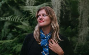 Mary Lattimore. A figure with dark, shoulder length blond hair looks to the left side. She is wearing red lipstick and a blue neck scarf. She is standing in a natural environment with ferns in the background.