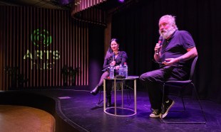 ArtsHub editors Thuy On and Richard Watts presenting the Crafting Insightful Criticism workshop at The Channel, Arts Centre Melbourne. Photo: ArtsHub. Two figures sit on stage beside a small coffee table with purple lighting. The figure on the left has black long hair and wearing a black dress with stockings and ankle boots. The figure on the right has grey short hair and a mid-length beard, wearing a black shirt, black pants and converse sneakers.