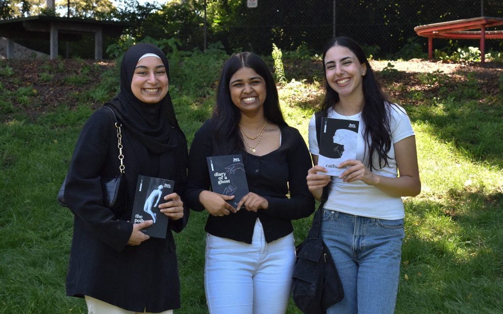 Young writers from Story Factory programs benefit in Create NSW’s Arts and Cultural Funding Program (ACFP). Photo: Aamina Musthafa. Three figures stand with a book in hand smiling towards the viewer with a green lawn behind them. The women on the left is wearing a black hijab and black long sleeve dress. The figure in the middle has black long hair and brown skin, wearing a black cardigan and light jeans. The figure on the right has light skin and dark brown long hair, wearing a white t shirt and jeans.
