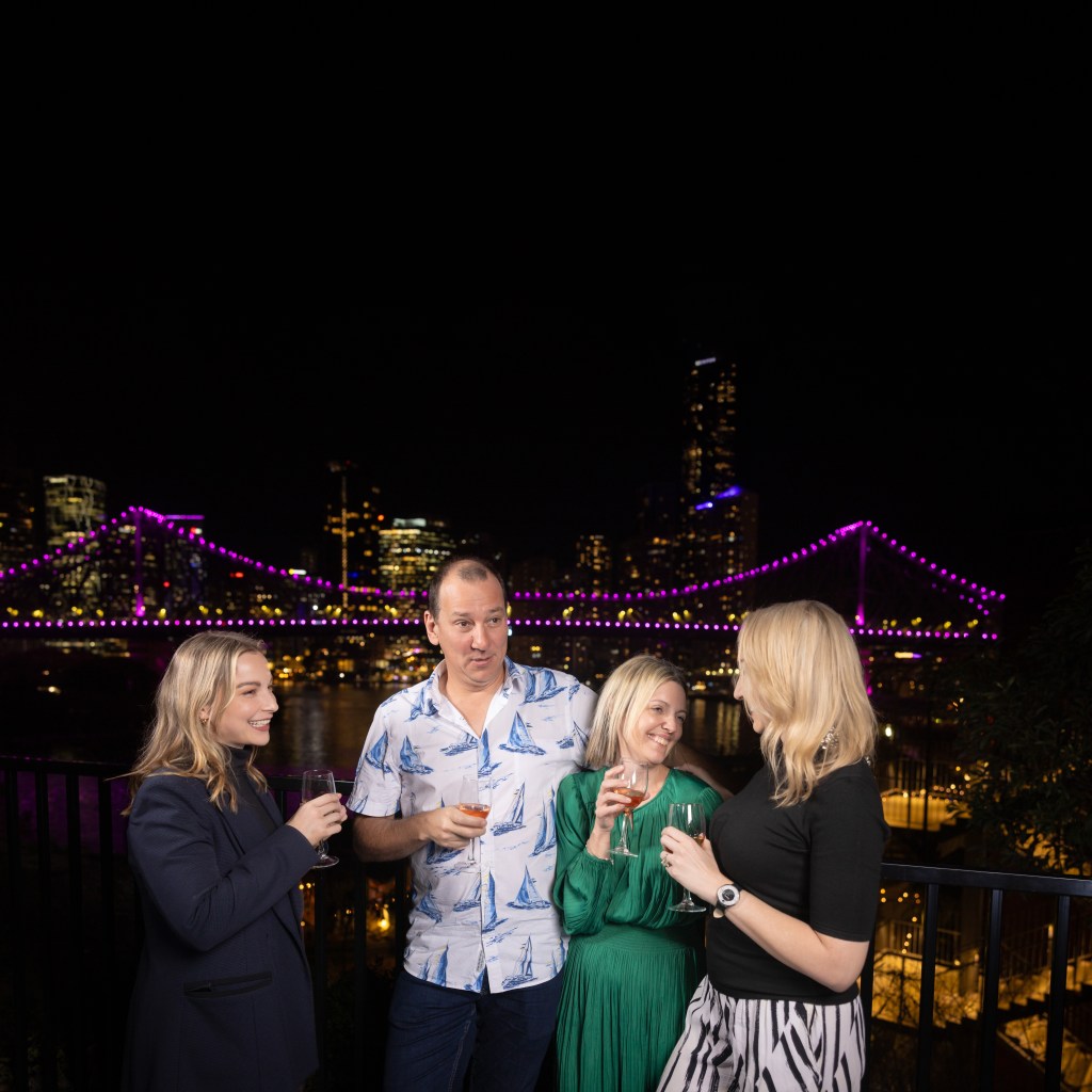 The cast of ‘All My Friends Are Returning to Brisbane’, (L-R) Sharnee Tones, Tim Jackman, Tammy Tresillian, Jaz Robertson. Photo: Kris Anderson. Four figures, three women with blond hair and a man with short brown hair, are having a glass of wine against a city night scape with colourful nights. 