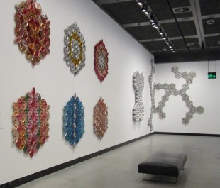 Donna Marcus mesmerises audiences with the mass and variety of vintage cookware she collected, sorted and assembled. 'Radiate' installation view at HOTA. Photo: ArtsHub. Red, blue and silver wall sculptures made of aluminium parts and arranged in different patterns arranged on a white wall.