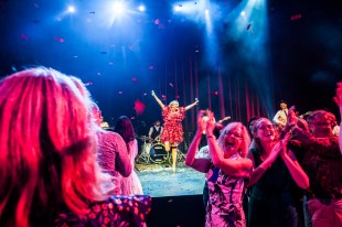 Christmas. A clapping and joyful standing audience looking around, while a performer with a red dress throws their arms in the air on the stage behind them.