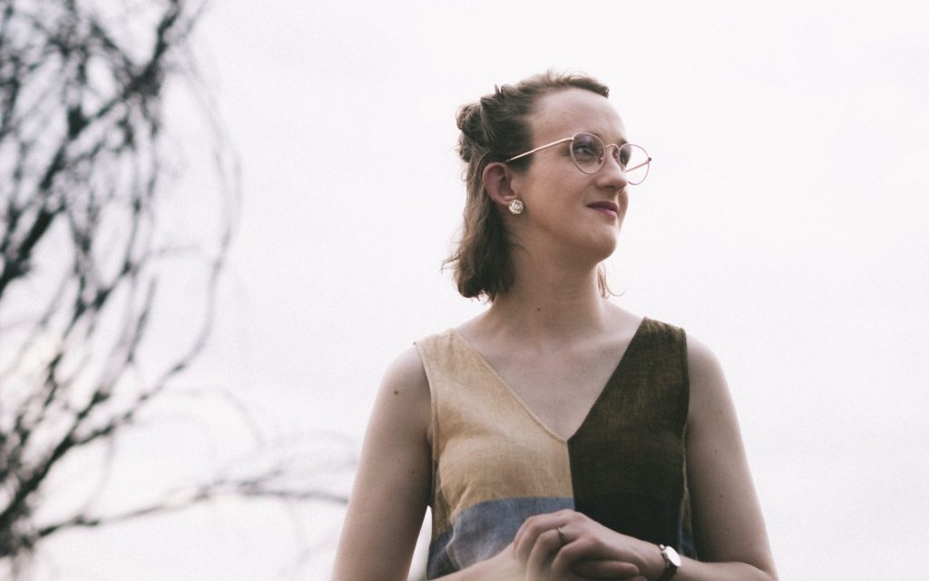 Melbourne Recital Centre welcomes Nat Bartsch as 2024 Artist-in-Residence. Photo: Supplied. Outdoor photo of a person with elegant short hair wearing glasses looking off into the distance with a smile, wearing a long patchwork dress.