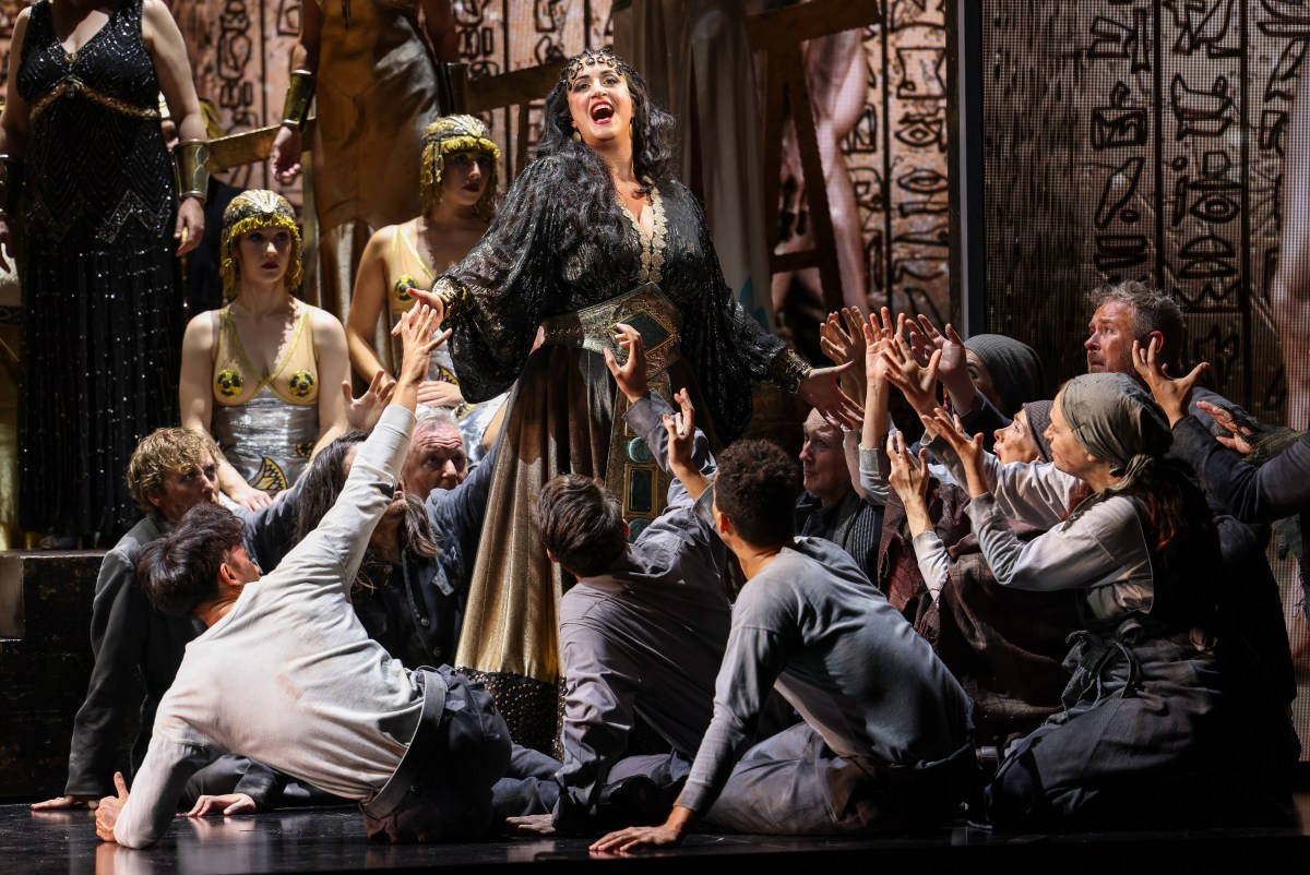 Aida. Image is a woman in black singing and gesticulating in operatic style, while members of the chorus lie around her reaching up, they are in Ancient Egyptian costumes, with lots of gold.