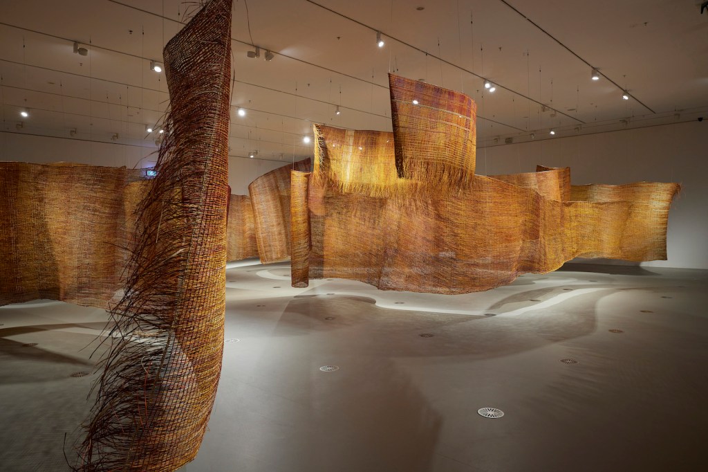 NGV Triennial. Image is huge collaborative sandy coloured weaving works handing from a gallery ceiling.