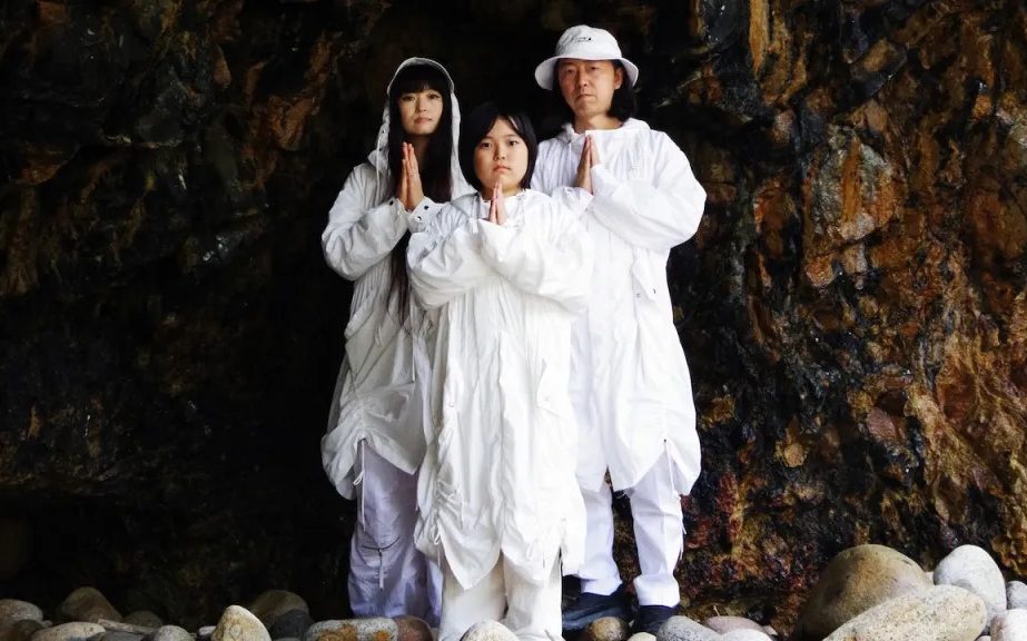 Families. Korean-Japanese ‘family’ band, Tennger. Photo: Supplied. Three figures stand in a triangle arrangement looking at the viewer with their hands in prayer. They are all wearing white flowing garments in a natural setting with a brown, rocky background.
