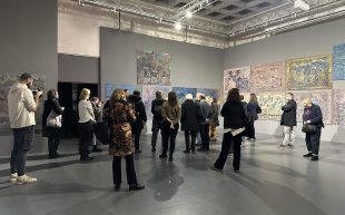 AICA members at the post-congress tour to BWA Tarnów. Photo: Lucy Hawthorne. A small crowd of people standing in front of artworks on a grey wall in an industrial looking space.