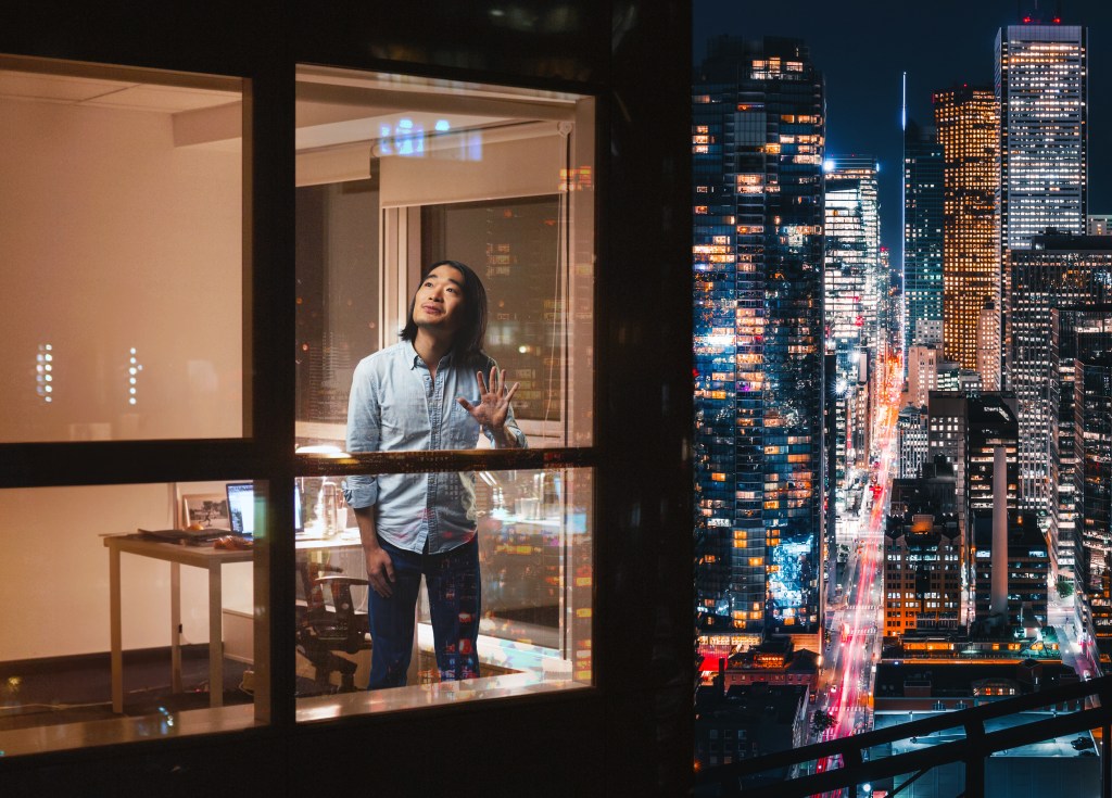 season. Image is a man in a high rise flat standing and looking out of the window.