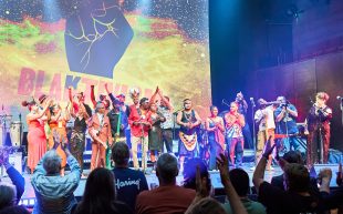 ‘BLAKTIVISM 2023’ performance at Hamer Hall, Arts Centre Melbourne. Photo: Pics That Pop. First Nations performers on stage wearing brightly coloured clothes against a projected background of a raised fist. In the foreground audiences are giving the performs a standing ovation.