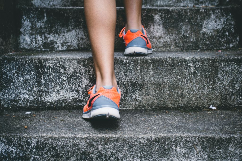 arts appointments. Close-up photo of a woman jogging up a flight of grey concrete steps. She is wearing orange and blue running shoes and has strong calves.