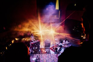 Phoenix. Image is a keyboard player, a drummer and a guitarist playing on a dark stage with two strong lights at the back.